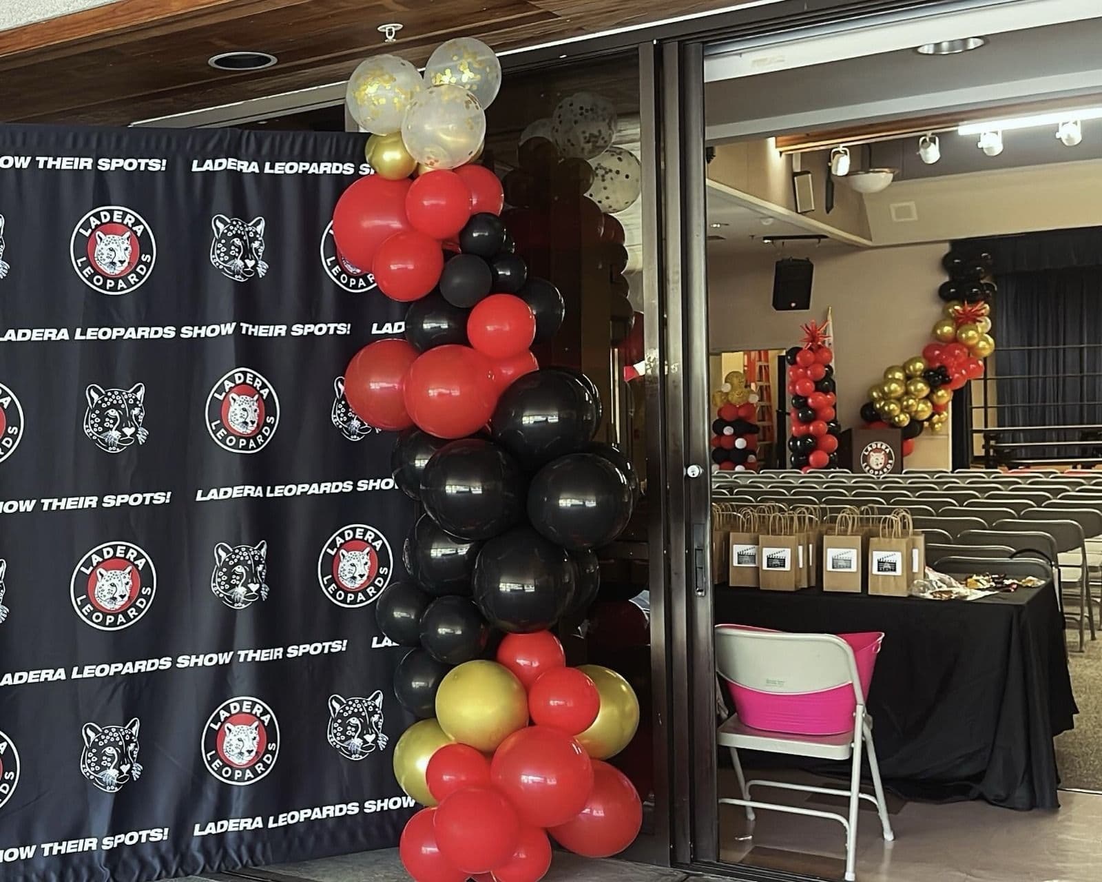 Red, black and gold balloon garland beside a team step-and-repeat banner at a hall entrance