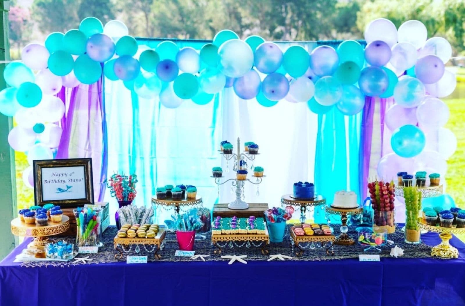 Turquoise and purple balloon garland over a birthday dessert table with a draped backdrop