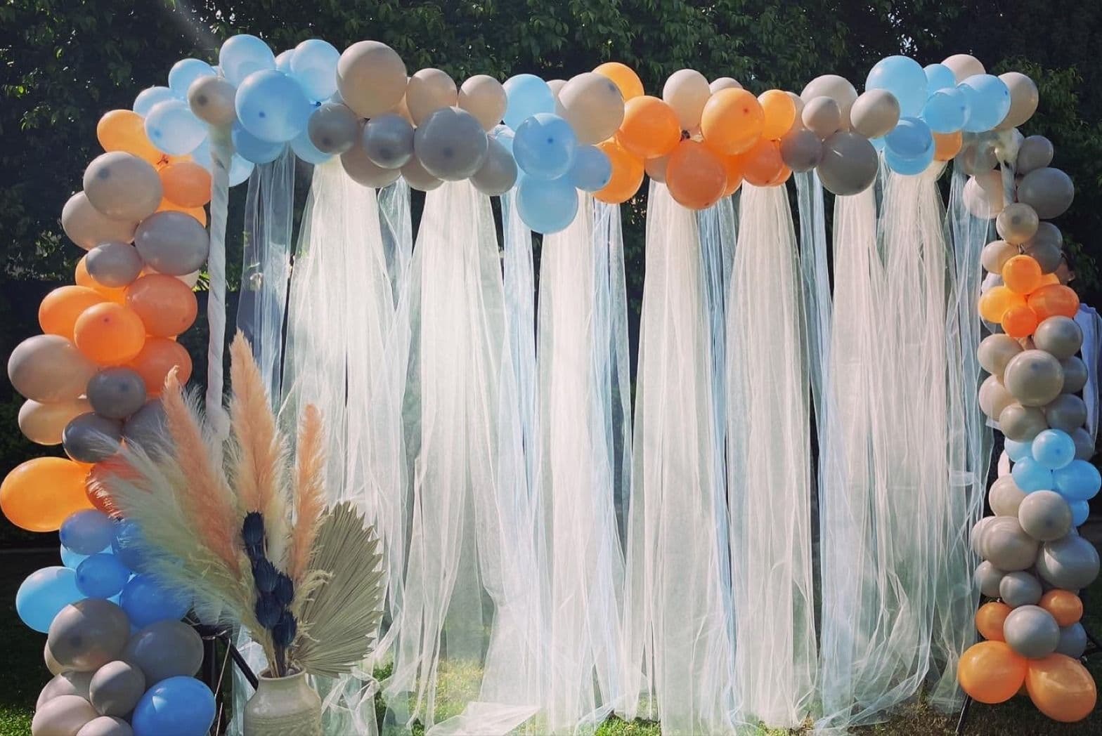 Blue, orange and grey balloon garland framing a white draped backdrop with pampas grass