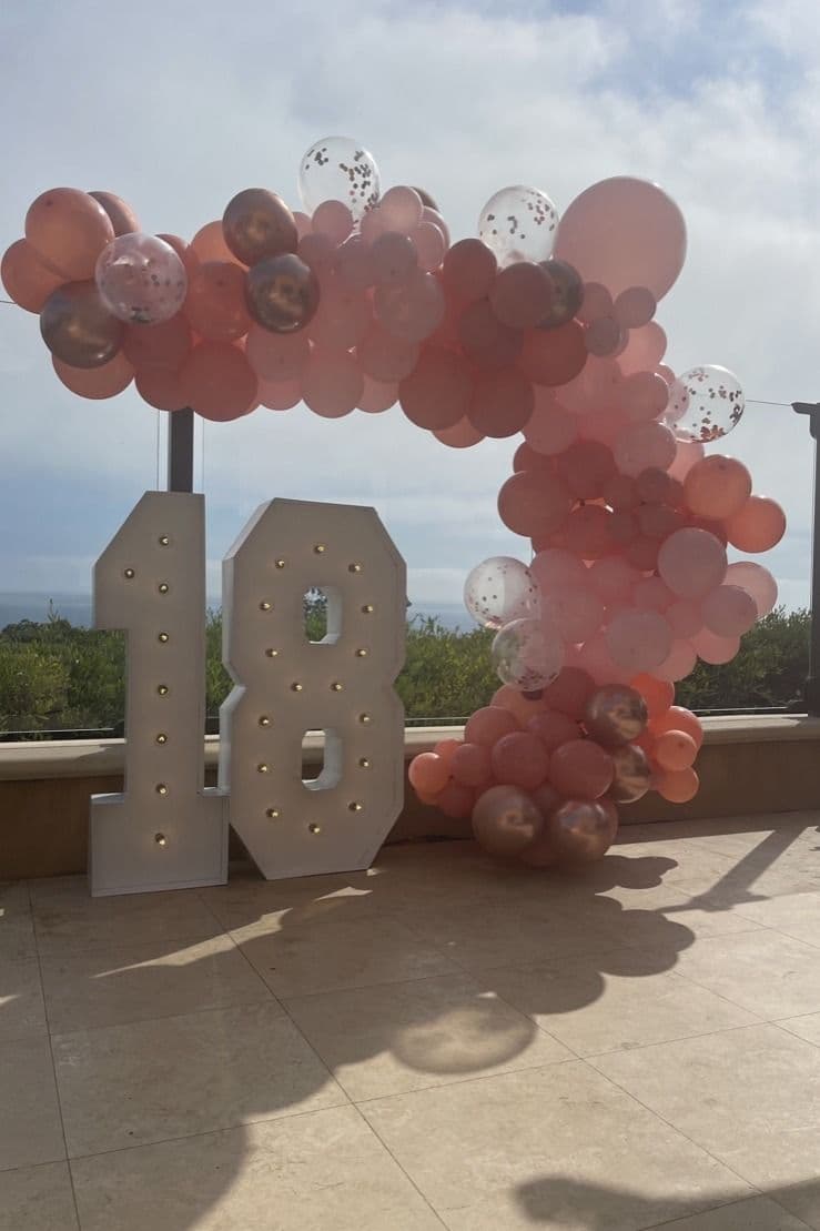 Rose gold and blush balloon arch over lit 18 marquee numbers on a terrace