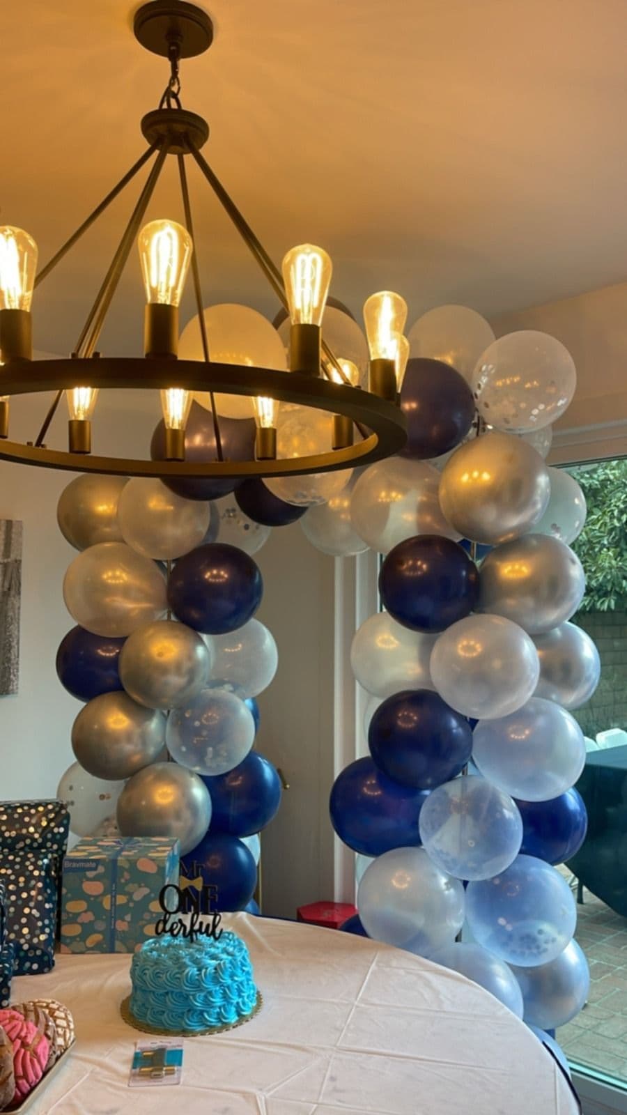 Navy, silver and gold balloon arch over a first birthday cake table beneath a chandelier