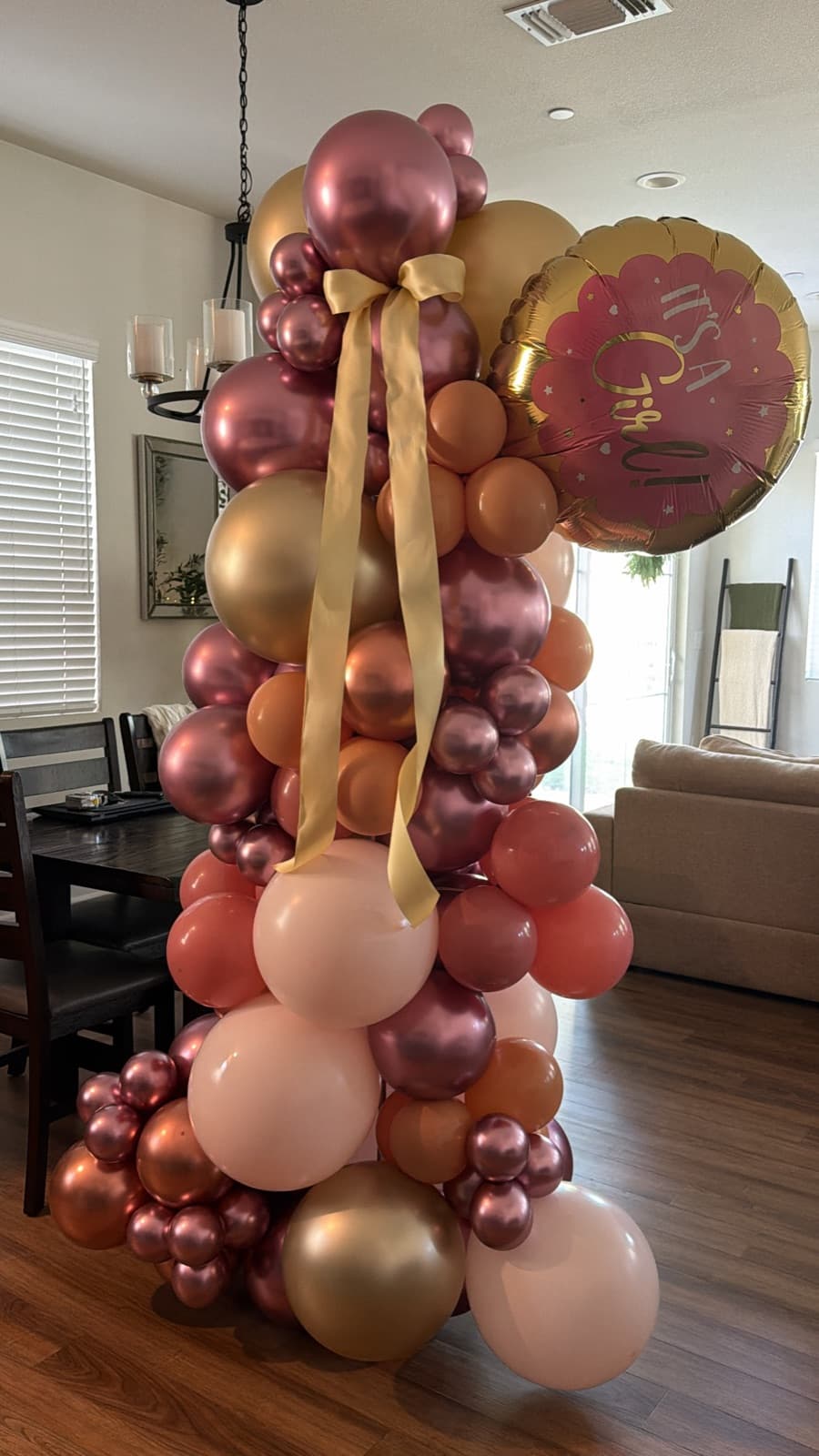 Rose gold, blush, coral and gold balloon column tied with a gold ribbon and topped with an It's a Girl foil balloon, in a living room
