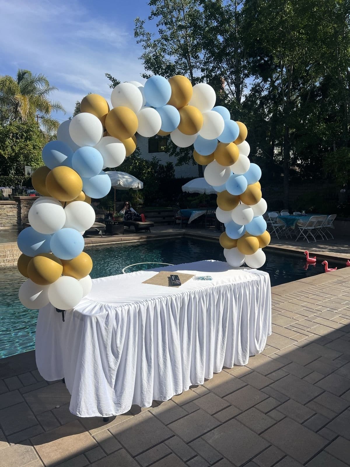 Blue, gold and white balloon arch over a draped table beside a swimming pool