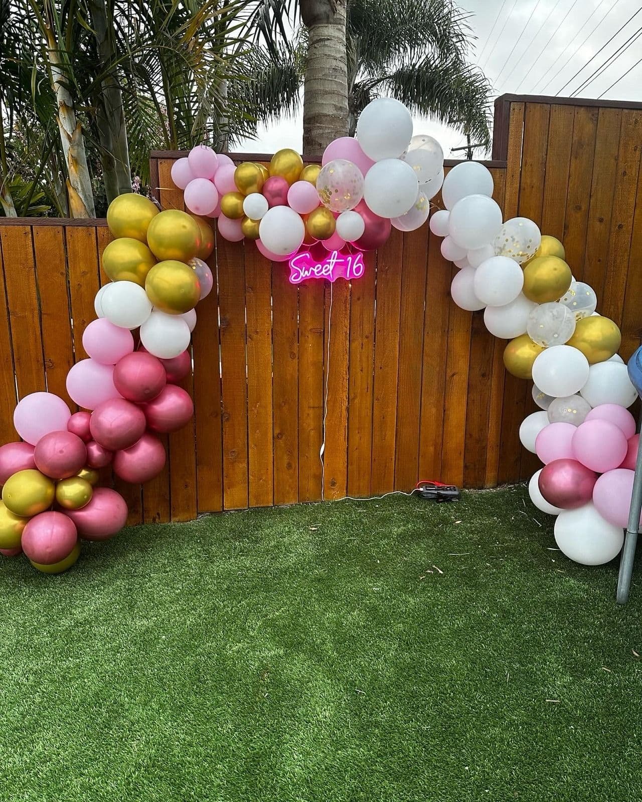 Pink, gold and white balloon garland along a wooden fence with a neon Sweet 16 sign