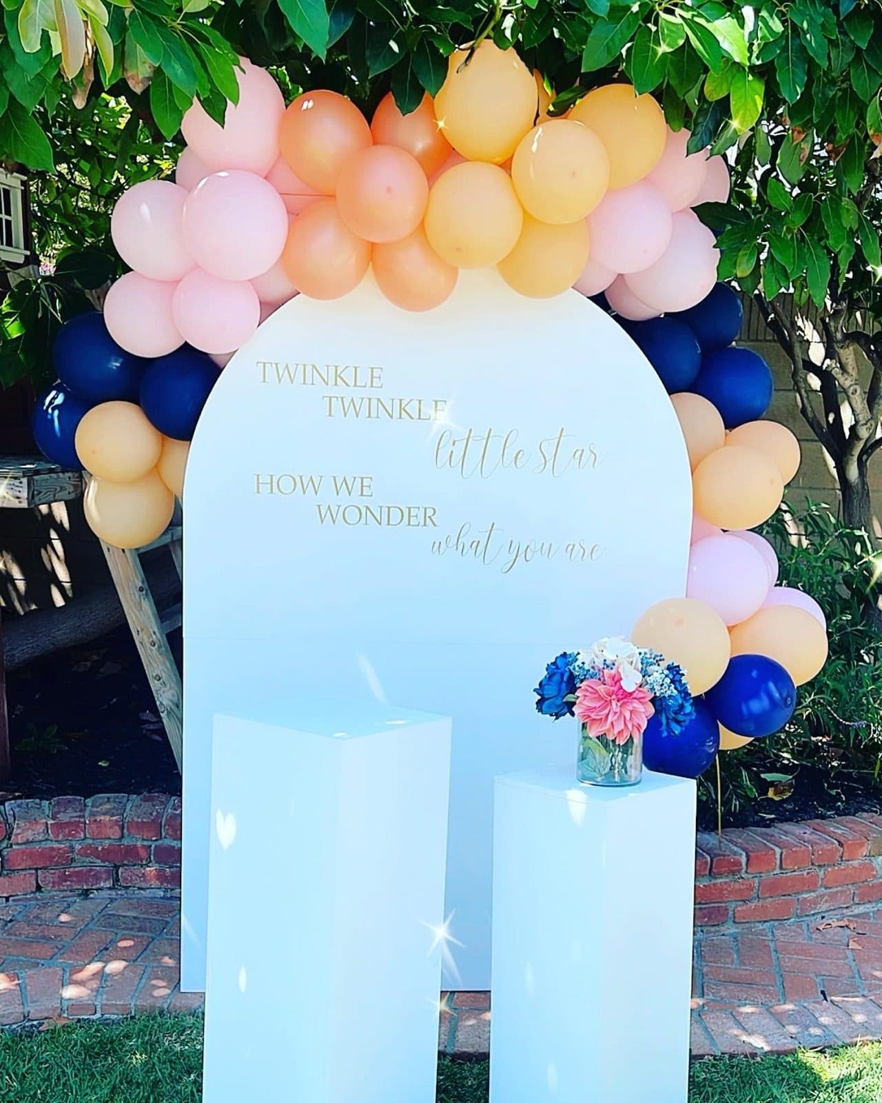 Peach, blush and navy balloon garland crowning a white arched backdrop in a garden